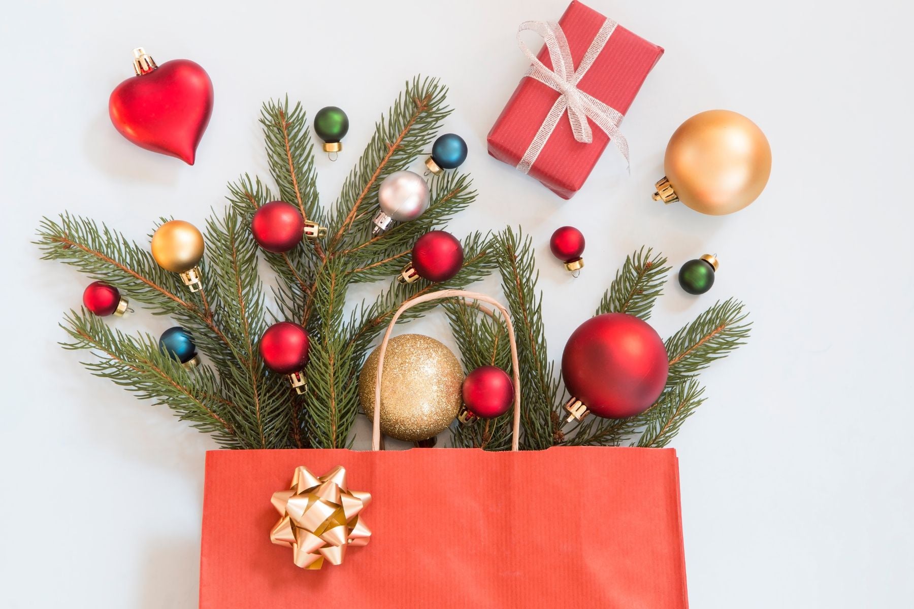Holiday shopping bag filled with pine branches, ornaments, and a wrapped box.