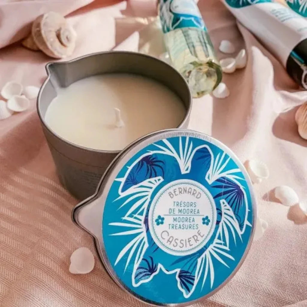 An open metal candle tin with creamy off-white candle wax beside its round lid, which has a blue tropical leaf design. The scene is styled on pink fabric with seashells and a matching bottle in the background.