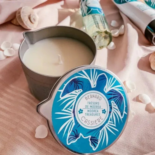 An open metal candle tin with creamy off-white candle wax beside its round lid, which has a blue tropical leaf design. The scene is styled on pink fabric with seashells and a matching bottle in the background.