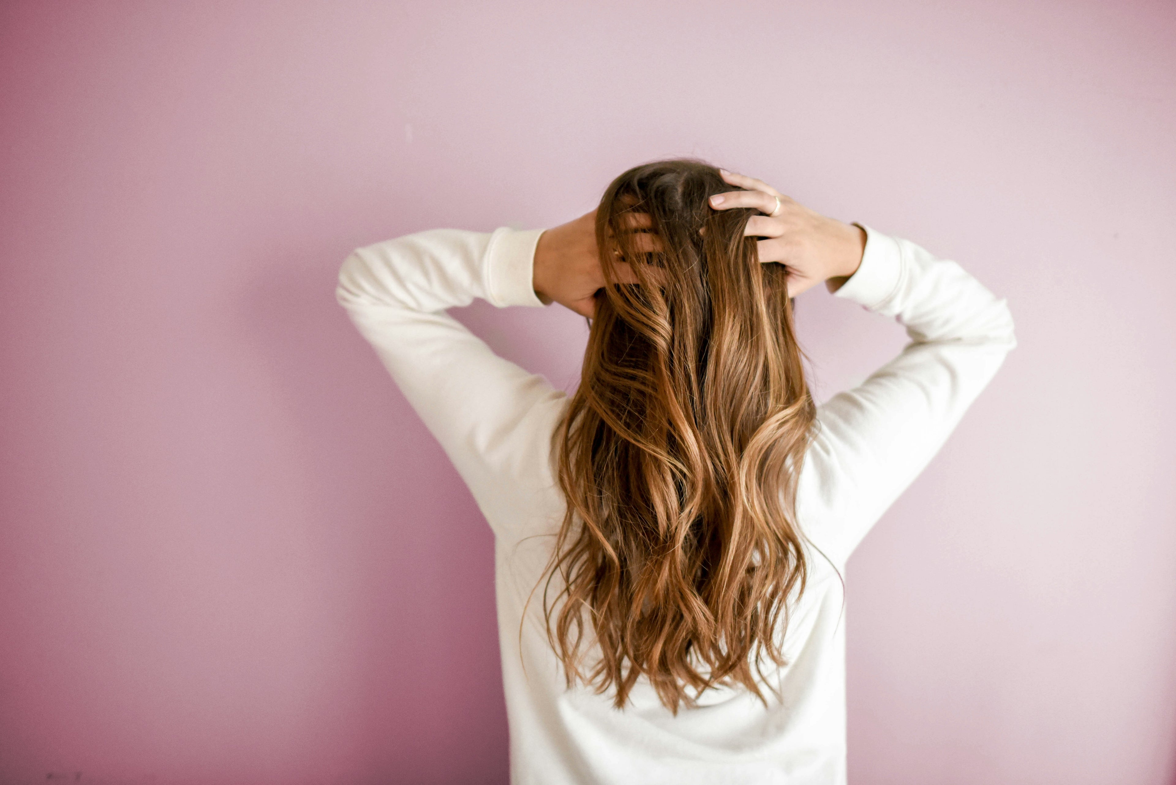 A person with healthy long hair touching their head against a pink background